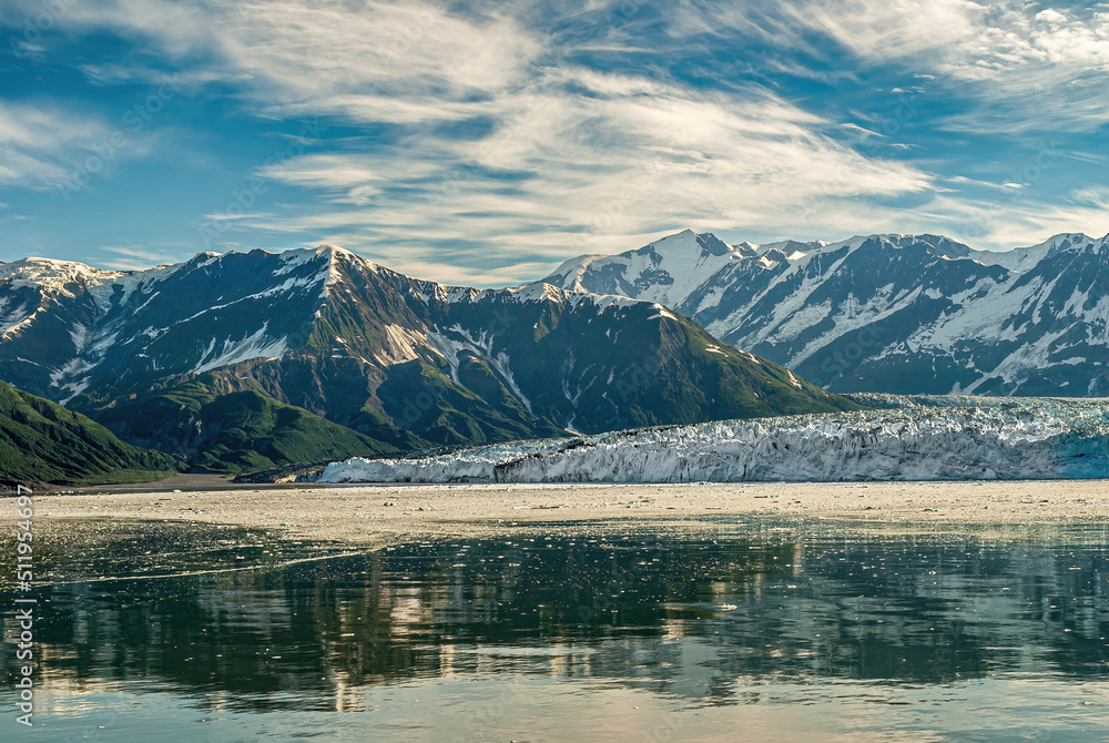 Disenchantment Bay, Alaska, USA - July 21, 2011: Hubbard glacier landing in ice covered ocean water with snow covered mountains in back under blue cloudscape. Green foliage on side.