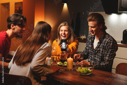 Joyful laughing woman showing funny selfie of her husband to couple of friends during double date dinner at home
