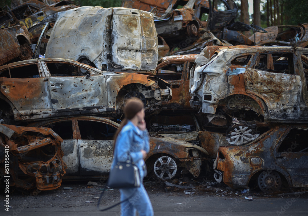 defocused girl talking on the phone walks past a pile of damaged cars in Irpin, Ukraine