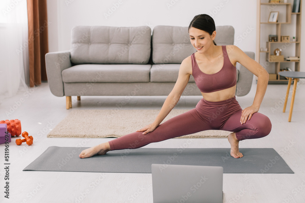 Fototapeta premium Sporty young woman practicing yoga in front of laptop, watching online tutorials.