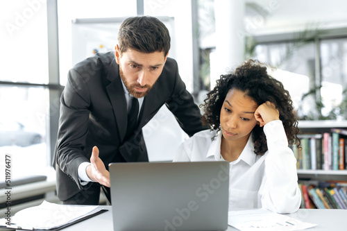 Upset focused multiracial colleagues, employees of an IT company, an African American woman and a Caucasian man, in a modern office, work on a laptop, look at the screen, test a new program © Kateryna
