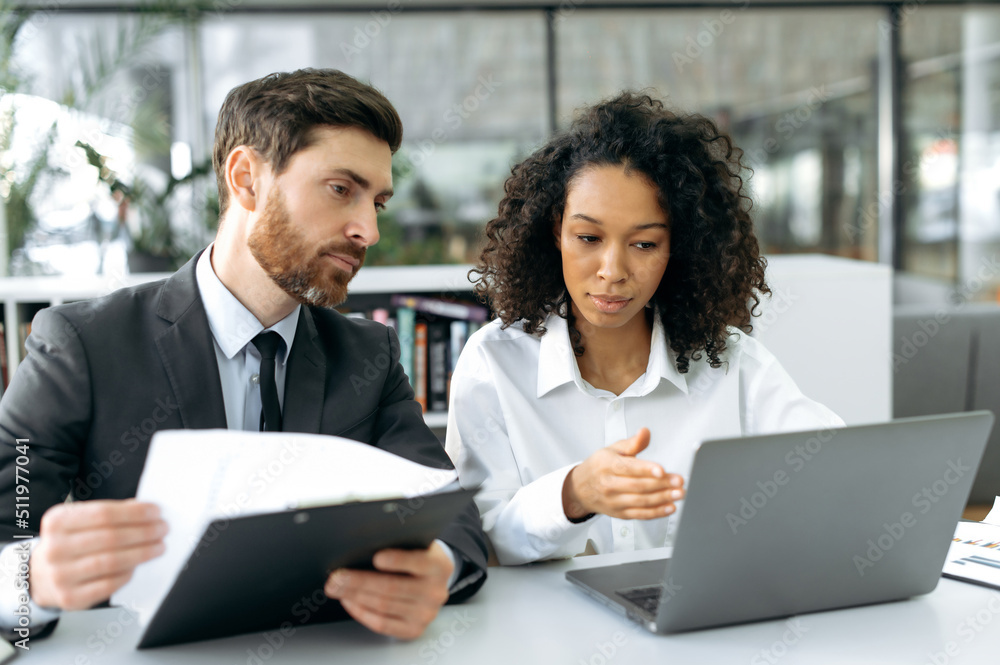 Business colleagues, an African American woman and a Caucasian man, company owners sitting at a desk in office, working on a laptop and with financial documents, preparing a contract for cooperation Business colleagues, an African American woman and a Caucasian man, company owners sitting at a desk in office, working on a laptop and with financial documents, preparing a contract for cooperation