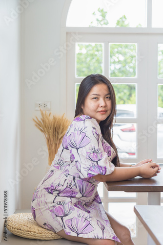 Portrait piceture of an Asian woman in a white patterned dress sitting and looking at a camera in a cafe