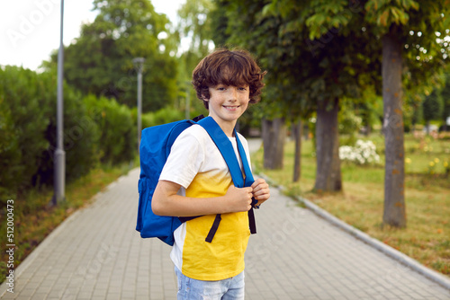 Portrait of cute preteen boy with school bag posing on warm morning on park walkway. Handsome caucasian little schoolboy with freckles wearing jeans and t-shirt smiling looking at camera. © Studio Romantic
