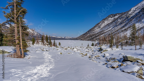Picturesque boulders are scattered in the snow-covered valley, coniferous trees grow. The path is trampled through snowdrifts. Ahead is a frozen lake surrounded by mountains. Altai. Multinskoye lake