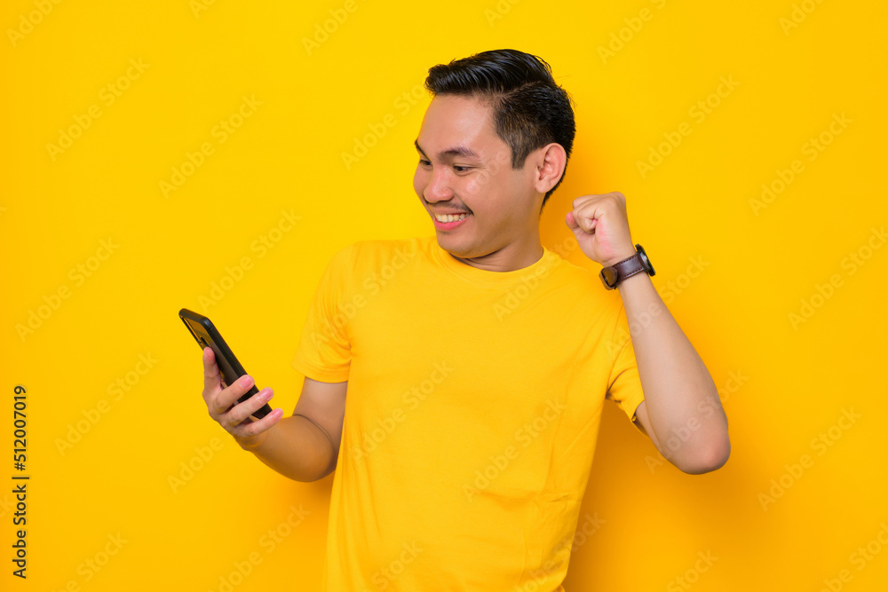 Excited young Asian man in casual t-shirt looking at mobile phone feel joyful reading good news isolated on yellow background. People lifestyle concept Excited young Asian man in casual t-shirt looking at mobile phone feel joyful reading good news isolated on yellow background. People lifestyle concept