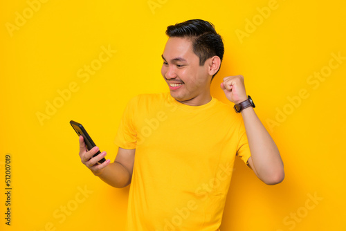 Excited young Asian man in casual t-shirt looking at mobile phone feel joyful reading good news isolated on yellow background. People lifestyle concept © Sewupari Studio