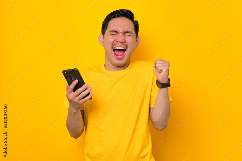 Excited young Asian man in casual t-shirt holding mobile phone and making winner gesture, getting good news isolated on yellow background. People lifestyle concept Excited young Asian man in casual t-shirt holding mobile phone and making winner gesture, getting good news isolated on yellow background. People lifestyle concept