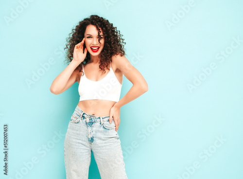 Sexy young beautiful smiling female in trendy summer hipster clothes. carefree woman posing near blue wall in studio. Positive brunette model with curls hairstyle. Cheerful and happy