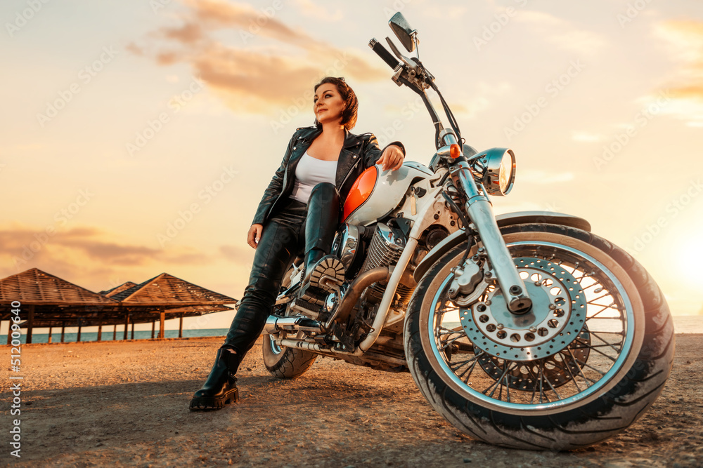 Obraz premium A beautiful independent woman in a leather clothes sitting on a motorcycle. Bottom view. Epic sunset on beach at the background. The concept of motorcycle trip and feminism