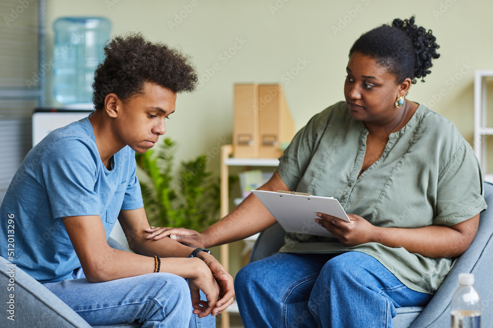African psychologist supporting depressed teenage boy feeling guilty about behavior during therapy at office African psychologist supporting depressed teenage boy feeling guilty about behavior during therapy at office
