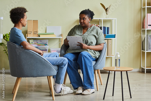 African school psychologist talking to difficult teenage boy during session while they sitting on armchairs at office photo