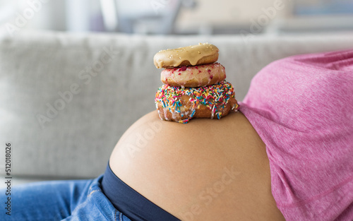 Pregnant woman with donuts on belly. Cravings of desserts and sweets during pregnancy, Girl eating unhealthy pastries on baby bump for gestational diabetes photo