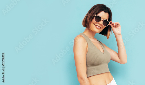Portrait of young beautiful smiling female in trendy summer clothes. Carefree woman posing near blue wall in studio. Sexy positive model having fun indoors. Cheerful and happy. Isolated. In sunglasses © halayalex