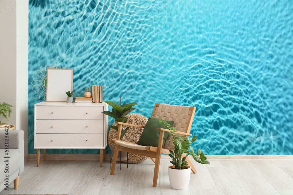 Comfortable armchair, chest of drawers and houseplants near wall with print of clear blue water