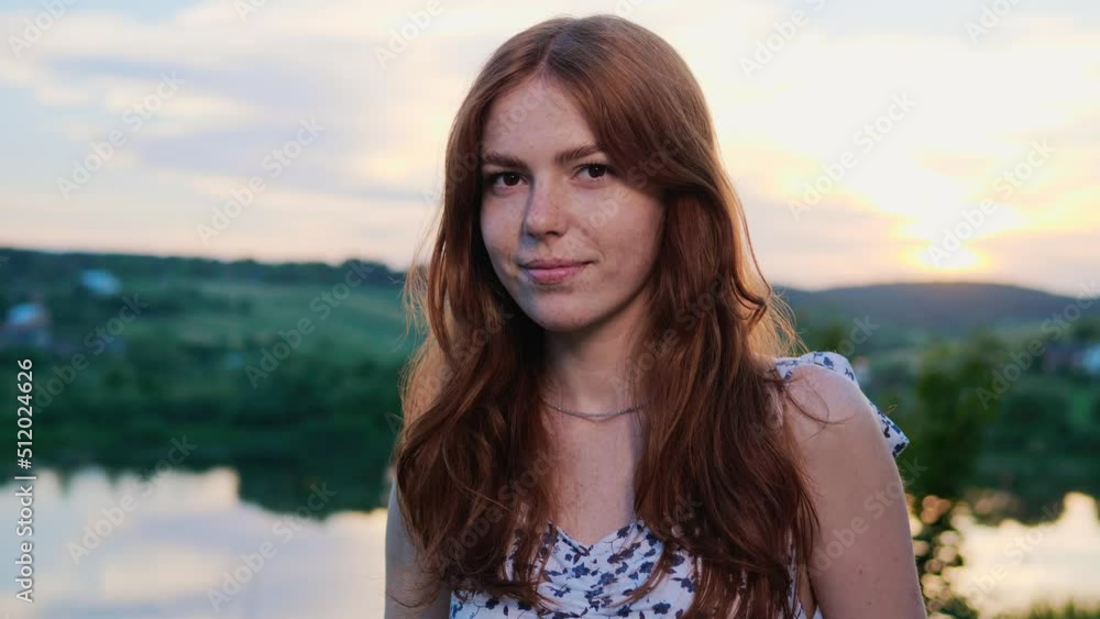 Pretty Happy Ginger Woman with long Red Hair Standing on background of the Lake on the Nature at Sunset, looking at Camera. Adorable Female Smiling Outdoor. Emotions. People. Portrait. Pretty Happy Ginger Woman with long Red Hair Standing on background of the Lake on the Nature at Sunset, looking at Camera. Adorable Female Smiling Outdoor. Emotions. People. Portrait.