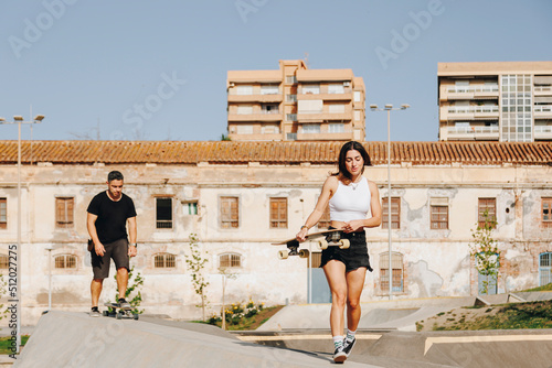 Young woman holding skateboard walking on sports ramp ahead of man photo