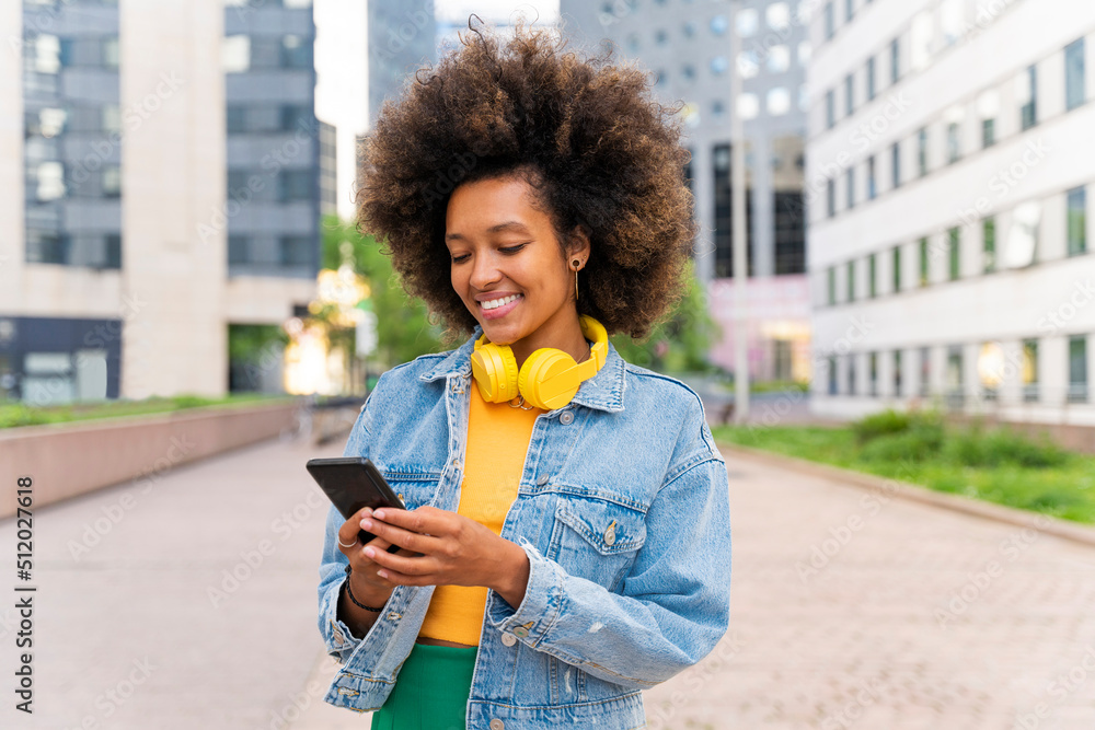 Smiling Afro woman text messaging through smart phone standing on footpath Smiling Afro woman text messaging through smart phone standing on footpath