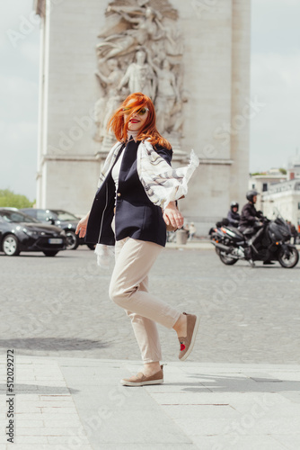 A woman in a navy blazer, cream trousers, wearing a white scarf and spinning in front of the Arc de Triomphe in Paris 