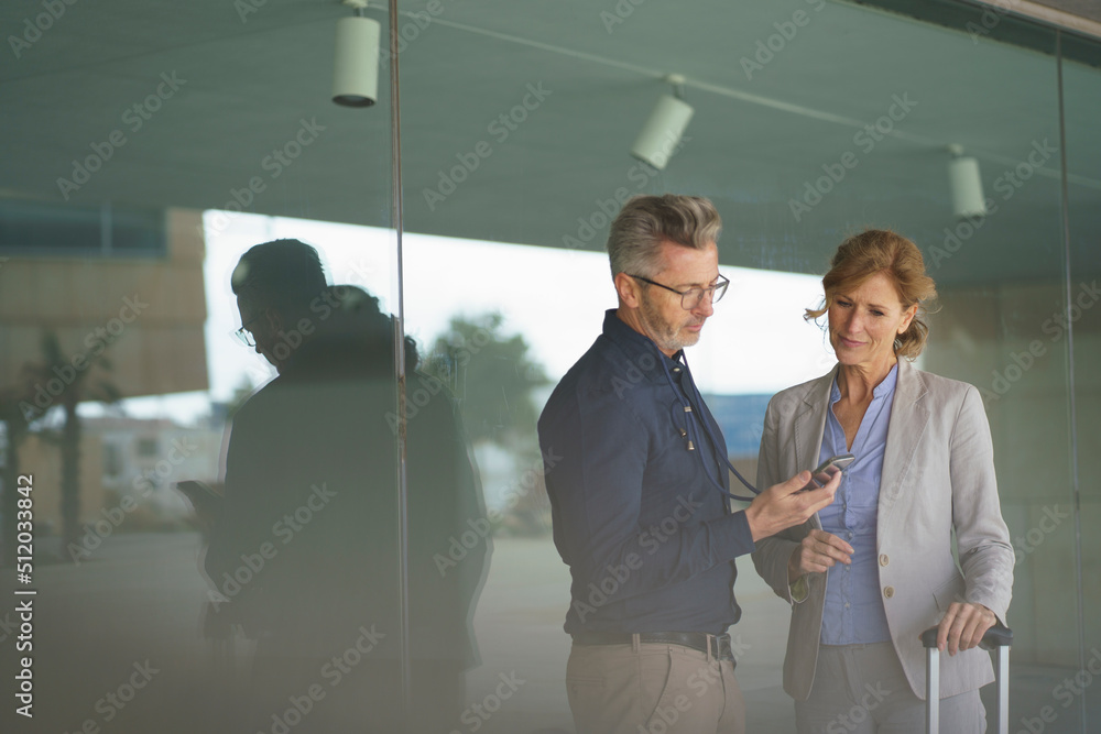 Businessman sharing mobile phone with businesswoman in front of glass wall Businessman sharing mobile phone with businesswoman in front of glass wall