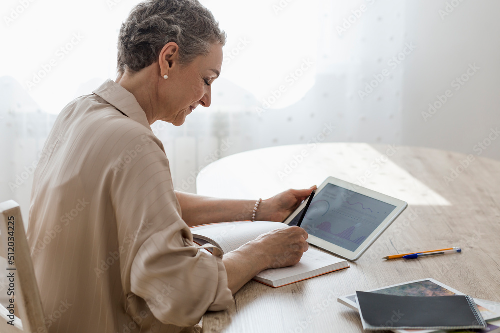 Woman holding digital tablet with data and taking notes at table at home Woman holding digital tablet with data and taking notes at table at home