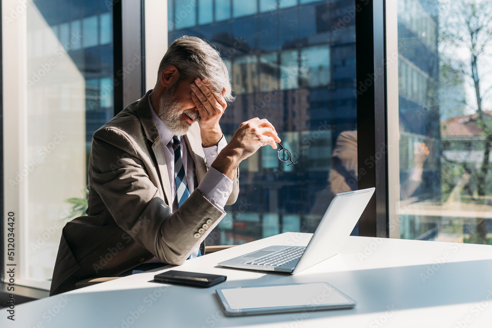 Tired businessman sitting with hand covering eye at desk in office Tired businessman sitting with hand covering eye at desk in office
