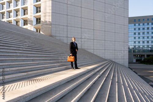 Mature businessman holding bag standing on staircase in front of building photo