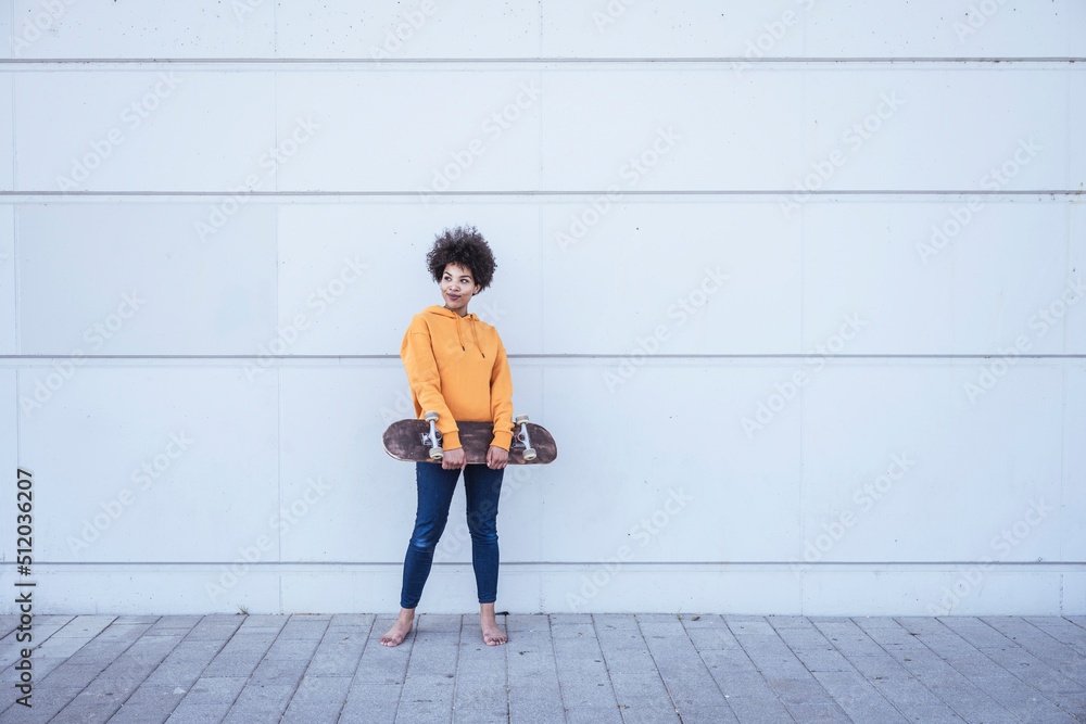 Young woman holding skateboard standing in front of wall Young woman holding skateboard standing in front of wall