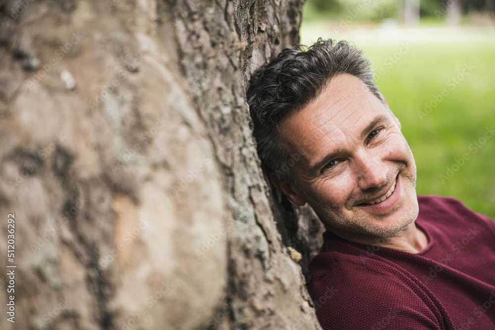 Smiling mature man leaning on tree trunk at park Smiling mature man leaning on tree trunk at park