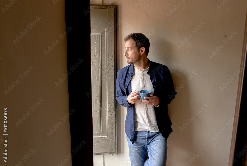 Thoughtful man with coffee cup leaning on wall looking out through window Thoughtful man with coffee cup leaning on wall looking out through window