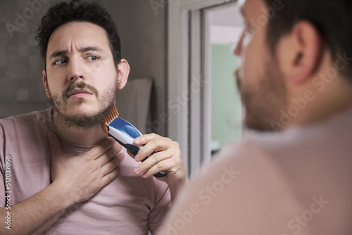 Young latin man looking at mirror and shaving beard with trimmer or electric shaver at bathroom.