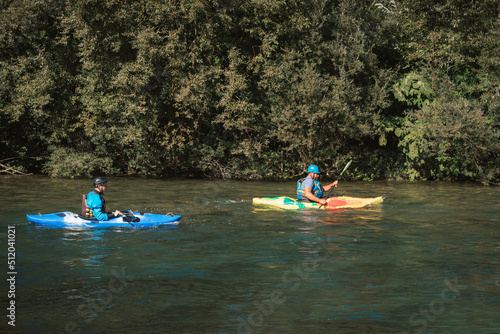 Two kayakers paddling and maneuvering on the fast-moving river with a rocky shore and going under the concrete bridge