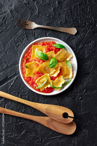 Ravioli with tomato sauce and fresh basil leaves on a plate, overhead flat lay shot on a black slate background with wooden spoons