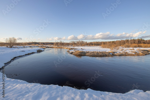March sunny evening by the river. Blue sky over the horizon. A picturesque landscape, early spring, a river with snow-covered banks, dry grass and bushes. The first thaws, the snow is melting © Sergei