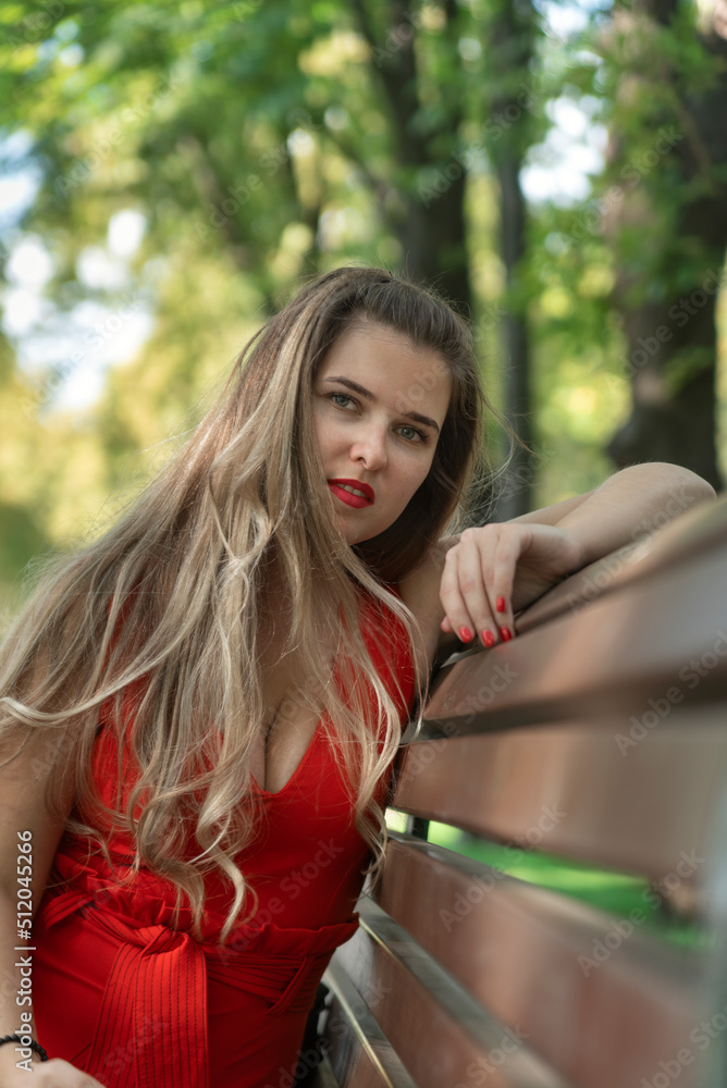 Naklejka premium Sexy young woman in red dress with long hair sitting on bench in park. Attractive girl. Vertical frame