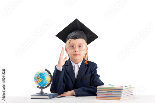 Serious boy first grader in students hat raising hand next to notebooks and globe. Studying and education concept.