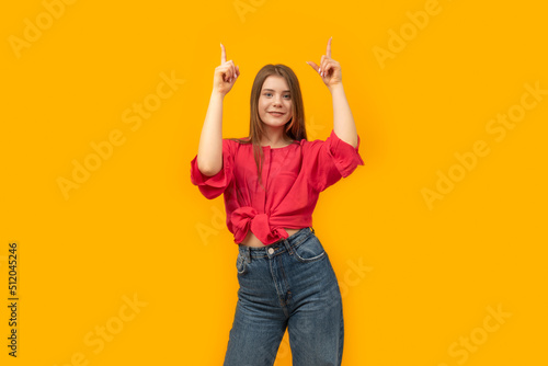 Attractive young woman looks straight and points her fingers up. Young smiling girl pointing up, yellow background