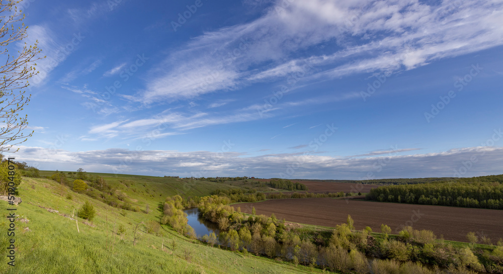 Landscape with a small river in a ravine. View of the countryside. bright greenery in the ravine. Saturated green grass against the blue sky. Plowed field on the horizon.