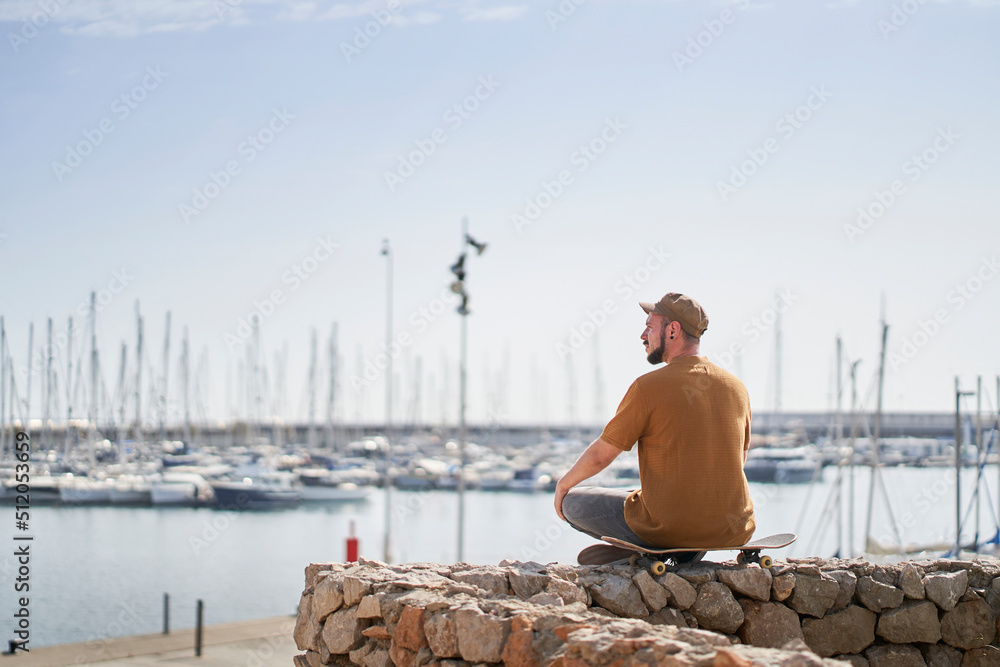 Man in cap relaxed sitting on a skateboard looking out over the harbor