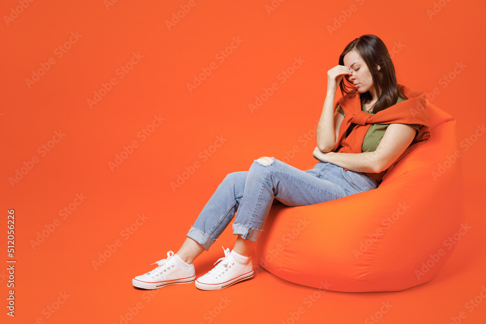 Full body young smiling happy woman 20s in khaki t-shirt sweater sit in bag chair keep eyes closed rub put hand on nose isolated on plain orange background studio portrait. People lifestyle concept. Full body young smiling happy woman 20s in khaki t-shirt sweater sit in bag chair keep eyes closed rub put hand on nose isolated on plain orange background studio portrait. People lifestyle concept.