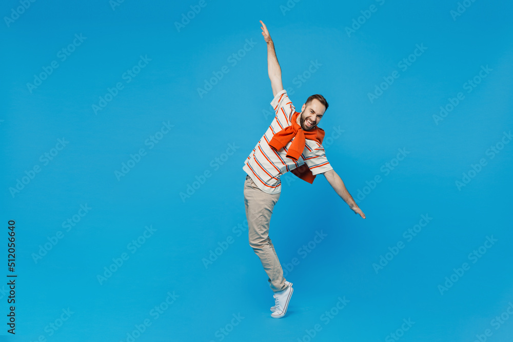 Full body young smiling fun man in orange striped t-shirt looking camera leaning back stand on toes dancing fooling around isolated on plain blue background studio portrait. People lifestyle concept.