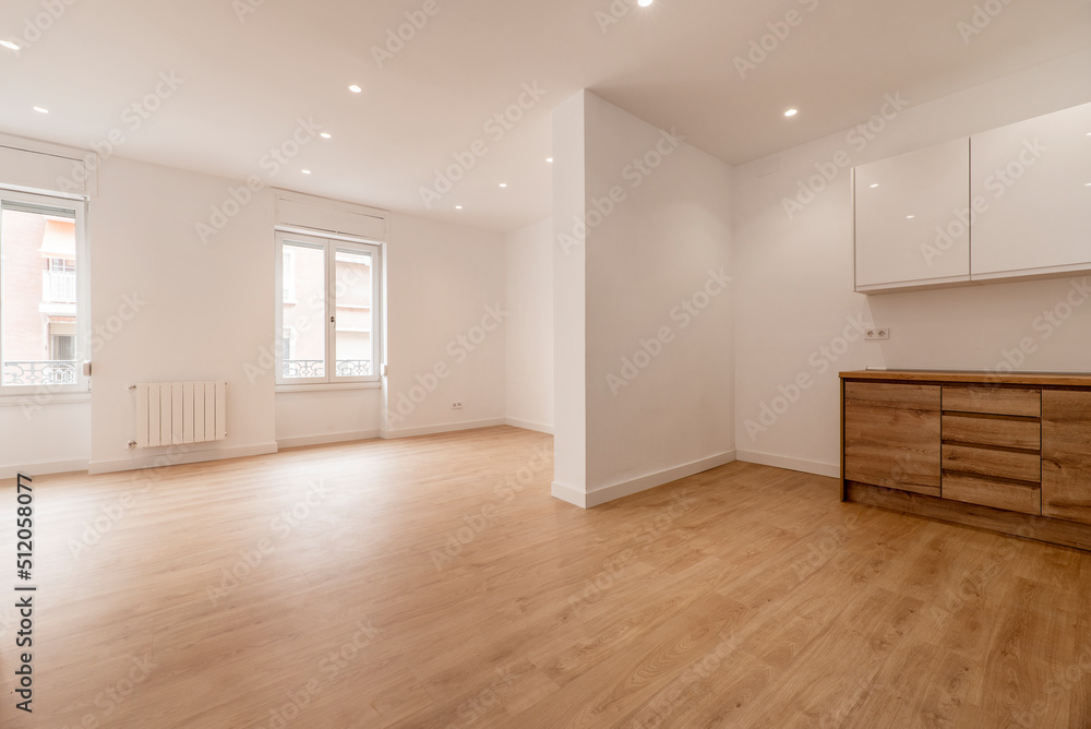 Empty living room with open kitchen with chestnut wood furniture, wooden floors and plain white painted walls Empty living room with open kitchen with chestnut wood furniture, wooden floors and plain white painted walls