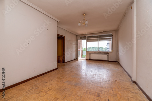 Empty room with white painted walls, oak parquet floor, white aluminum radiator under the large aluminum window of a terrace © Toyakisfoto.photos