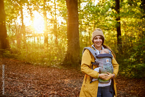 Portrait of smiling mother carrying sleeping baby in baby carrier in woodland, autumn photo