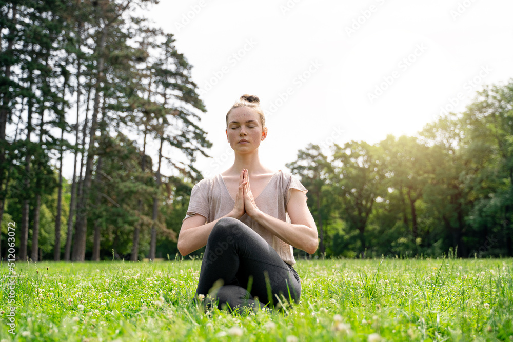 Young caucasian slim fit blond hair carefree calm woman sitting in lotus yoga pose with closed eyes and hands as in prayer, meditating on green grass meadow in summer park on early morning or sunset Young caucasian slim fit blond hair carefree calm woman sitting in lotus yoga pose with closed eyes and hands as in prayer, meditating on green grass meadow in summer park on early morning or sunset