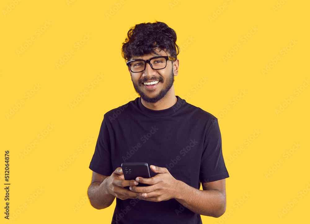 Crop shot of smiling millennial Indian guy in glasses isolated on yellow studio background using modern smartphone. Happy young biracial man text or message online on cellphone. Communication. Crop shot of smiling millennial Indian guy in glasses isolated on yellow studio background using modern smartphone. Happy young biracial man text or message online on cellphone. Communication.