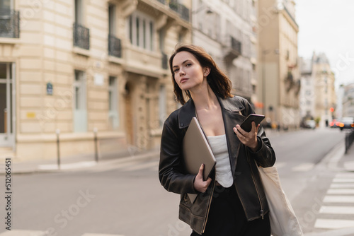 stylish freelancer in black leather jacket holding laptop and smartphone on street in paris.