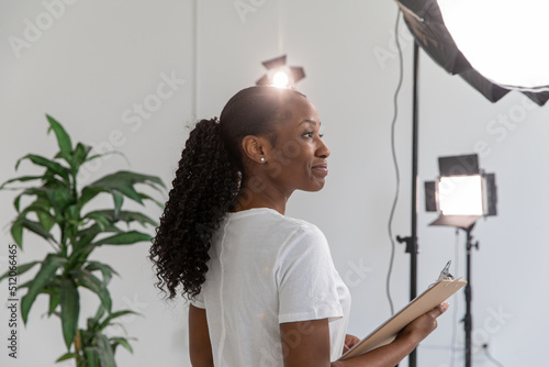 African American Woman Working on Film Set Holding Clipboard With Lighting and Equipment in Background. Proud Female Director in Video Production Studio