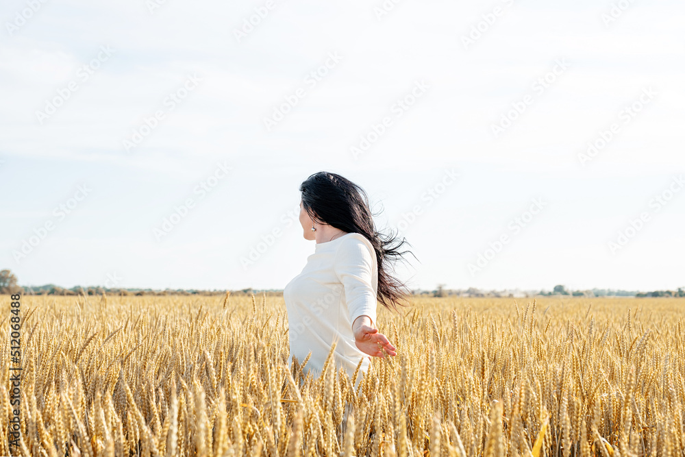 Mid adult woman in white dress standing on a wheat field with sunrise on the background, back view Mid adult woman in white dress standing on a wheat field with sunrise on the background, back view