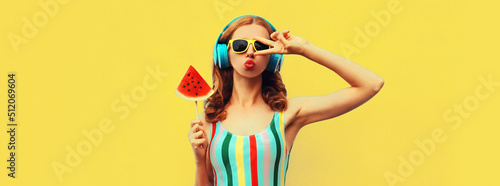 Summer portrait of stylish woman in headphones listening to music blowing her lips with juicy lollipop or ice cream shaped slice watermelon on yellow background, blank copy space for advertising text